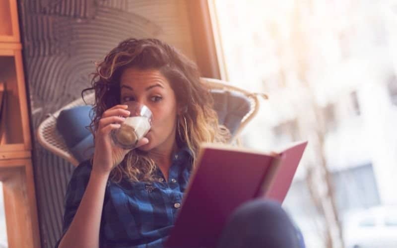 a woman sitting on a chair while holding a book she is reading and at the same time drinking a beverage from a glass that she is holding in another hand