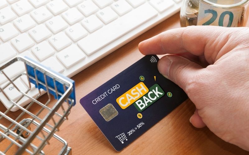 Photo of a hand holding a cashback card while in front of a laptop beside a toy grocery cart