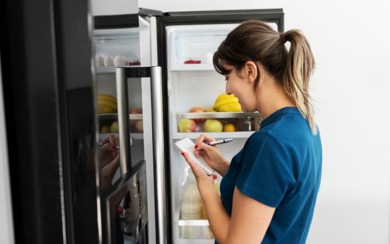 Photo of a woman in front of an openned refrigerator while making a list