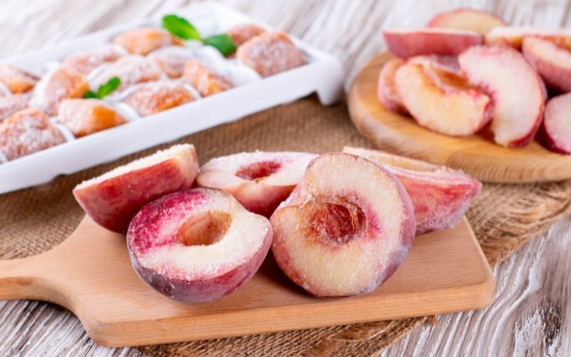 Photo of frozen sliced apples and pears on a wooden board beside a white tray filled with other kinds of frozen fruits