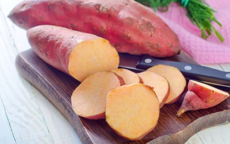 Photo of whole and sliced sweet potatoes with aknife between then on a wooden chopping board