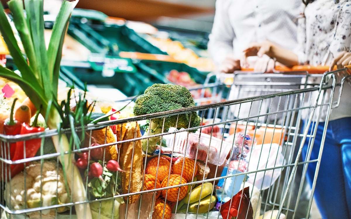 Photo of a grocery cart filled with vegetables, fruits and other items pushed by a woman