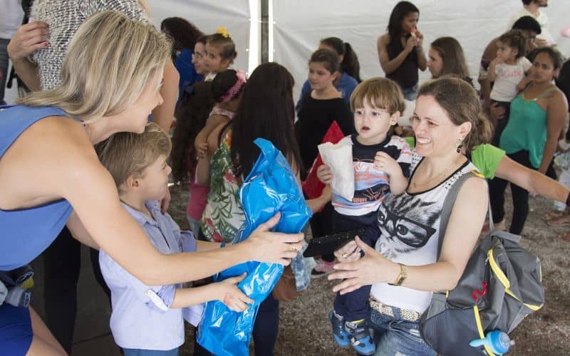 A covered area full of people including a woman and a girl giving something wrapped in blue plastic wrapper giving it to a woman carrying a kid