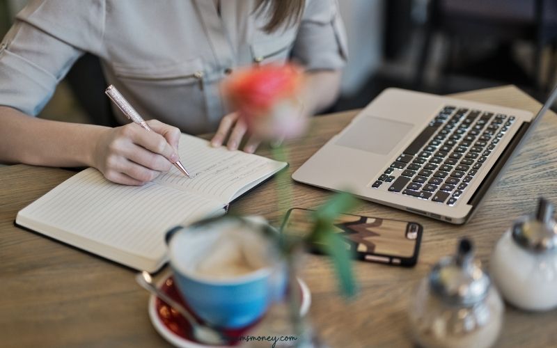 woman writing in notebook with coffee and laptop on table at a cafe