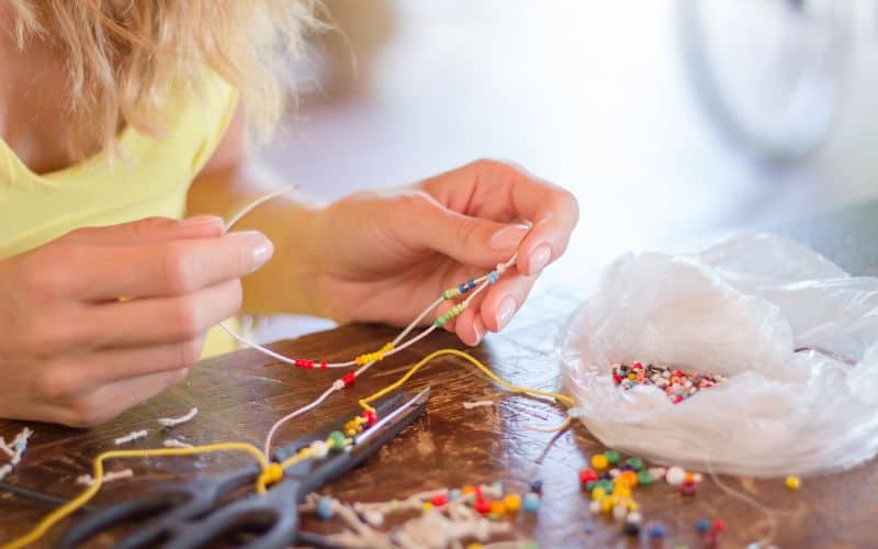 a pair of hands holding a thread with beads and more small colorful beads and a scissor on the table 