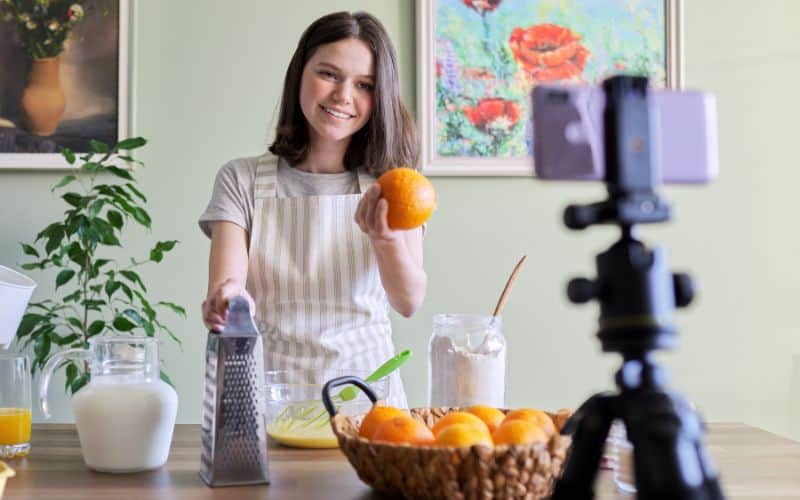 A young woman holding an orange with more orange on a table while in front of a camera for the you tube to make money