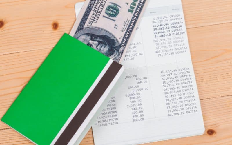 Photo showing a green booklet, some money and an open booklet with numbers on wooden table