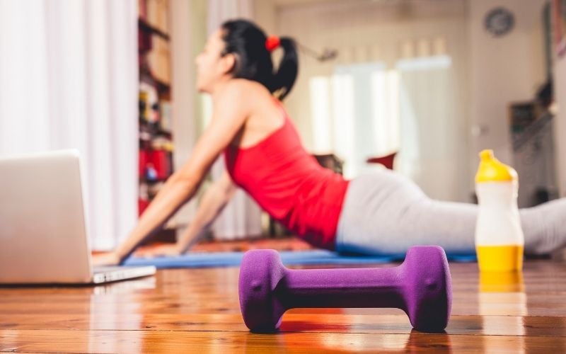 Photo of a woman on the floor doing a yoga en excercise with a dumbbell and a laptop nearby