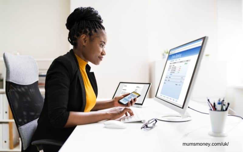 woman with yellow top sitting at desk holding smartphone looking at computer