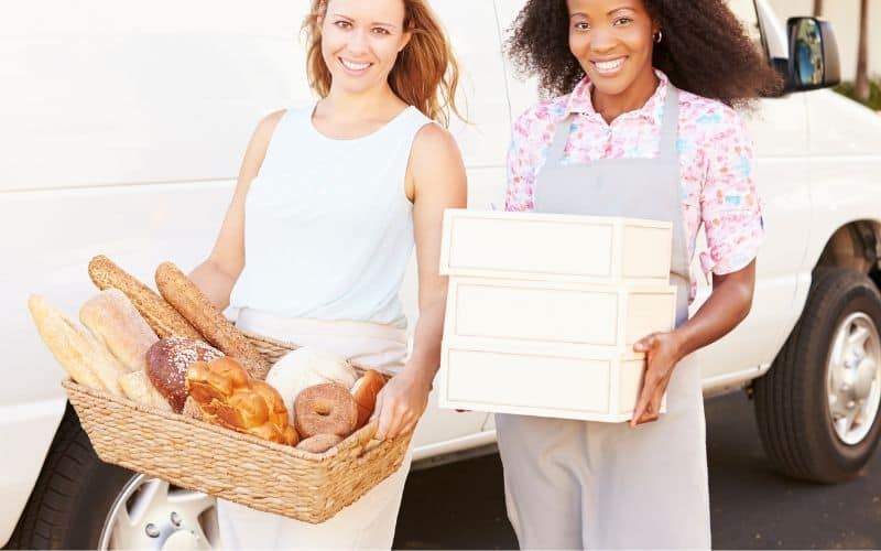 Two women with one holding a bax of different bread while the other is holding 3 white boxes beside a white van