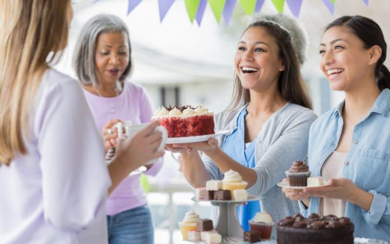 A photo showing two happy women offering cakes they are selling to the two women in front of them with more cakes on the table.