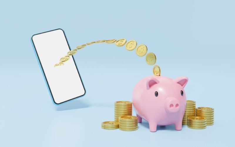 Image of a cellular phone with coins around the pink piggybank in blue background