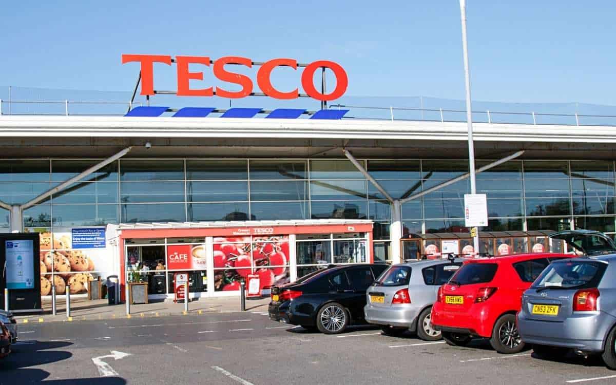 A large supermarket with the word Tesco sign above the entrance and parking lot with cars at the front