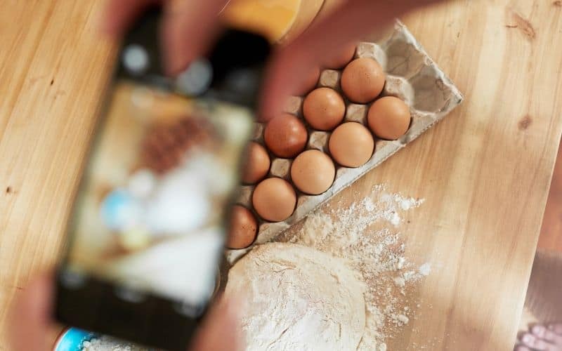 Image showing a hand holding a smart phone and taking picture of a tray of eggs with flour beside it.