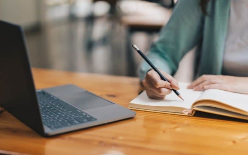 Photo of a hand holding a pen while writing on a notebook in fron t of a laptop.