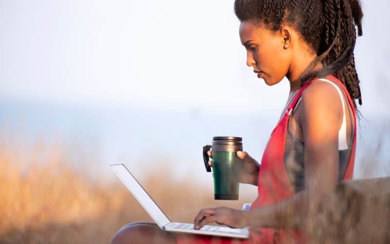 Photo of a woman sitting in a grassy area holding a coffee mug typing on a laptop on her lap.