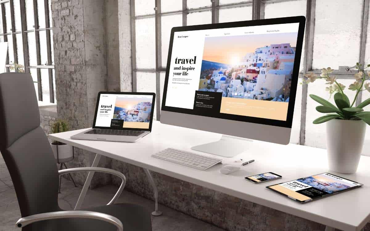 An image showing computer monitor, a laptop, a smart cellphone and a tablet on white table with swivel chair beside it showing a website.