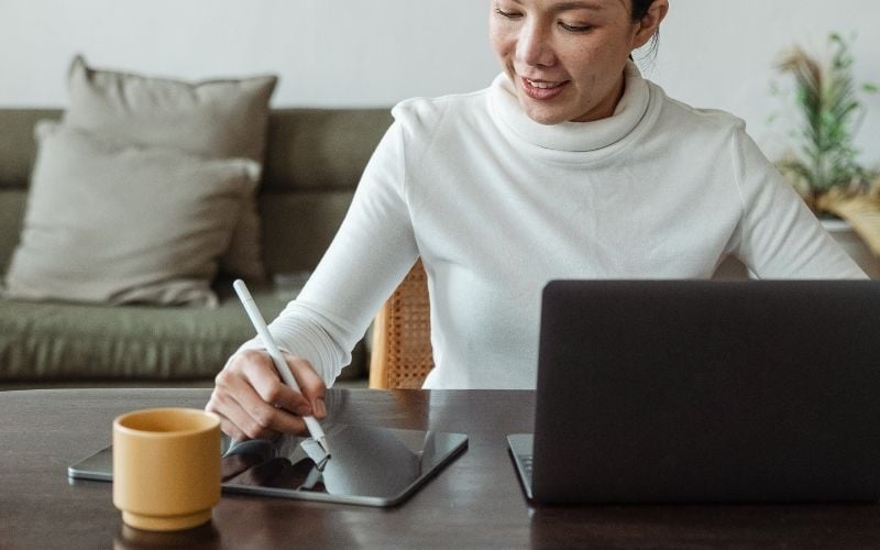 Photo of a woman holding a pen writing on a tablet and in front of a laptop